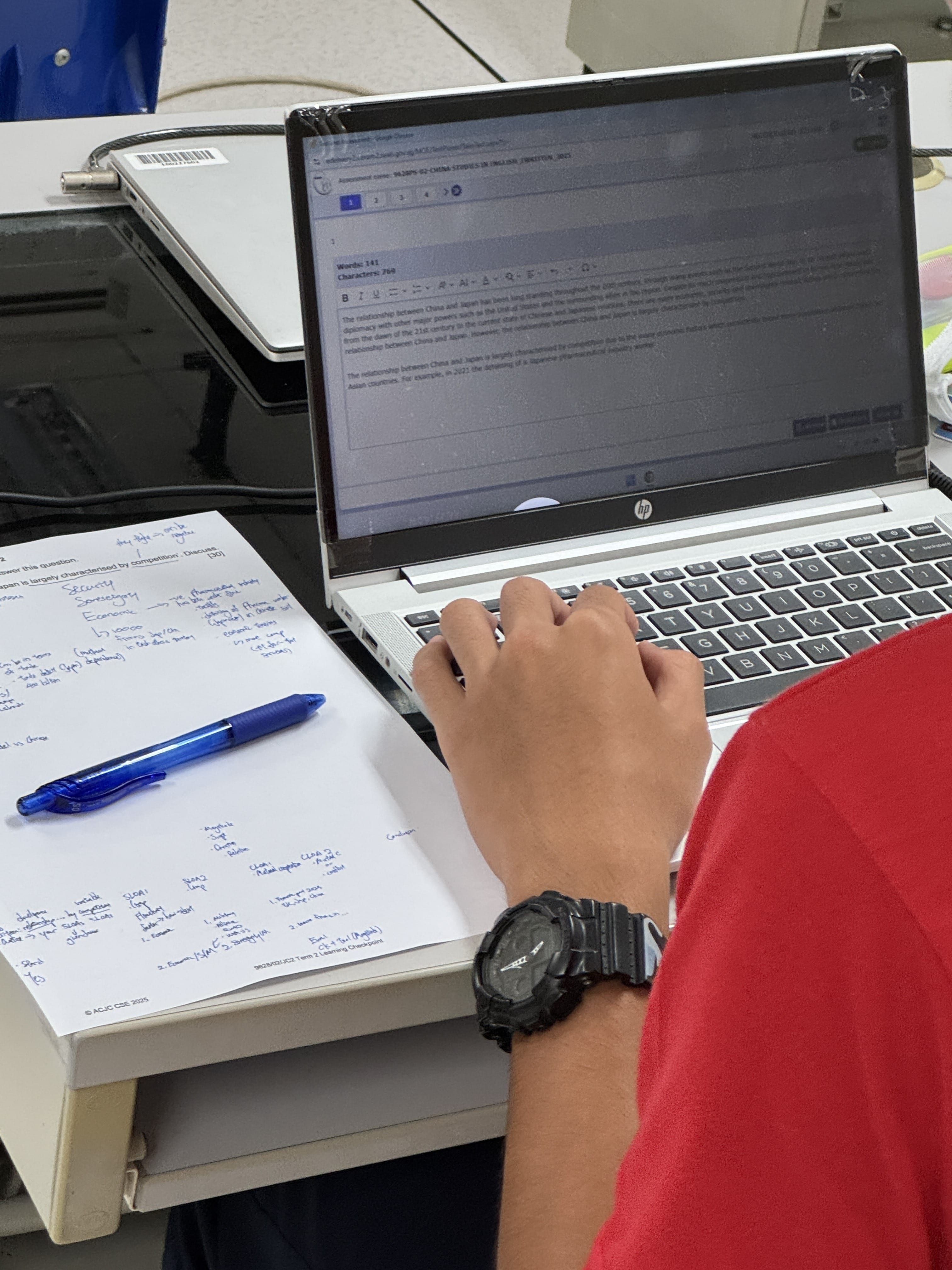 A person wearing a red shirt types on a laptop beside a sheet of handwritten notes and a blue pen.